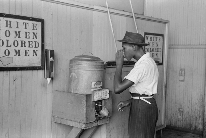 A photograph titled “Negro drinking at ‘Colored’ water cooler in streetcar terminal, Oklahoma City, Oklahoma,” by Russell Lee (1903–1986). This photo captures the ways in which Jim Crow laws, which enforced racial segregation in the United States, affected the everyday lives of Black Americans, who were required to use different services than White Americans used.
